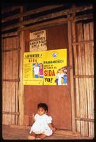 A baby sits under signs for Panamanian AIDS prevention, Darien Gap, Panama